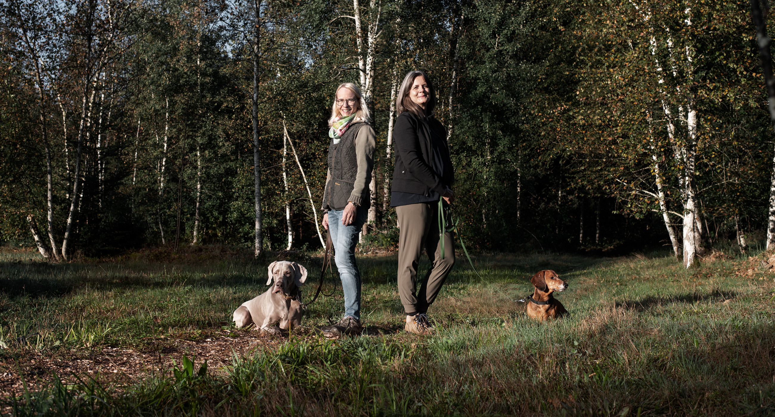Christine Müller und  Nicole Bernhardsgrütter stehen mit ihren Hunden Jury und Rufus auf einer sonnigen Waldlichtung