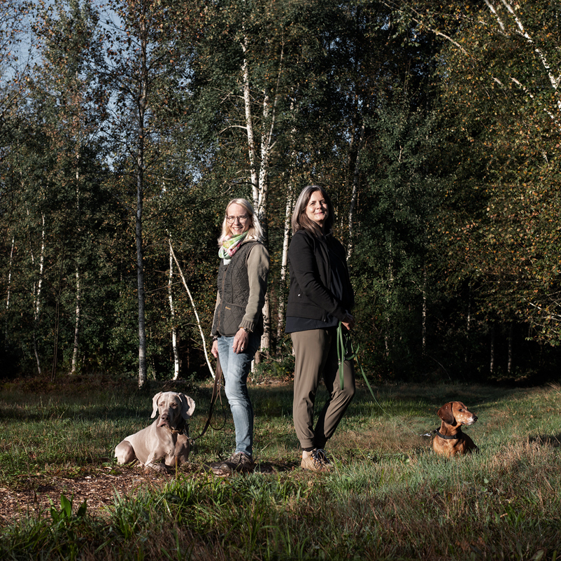 Christine Müller und  Nicole Bernhardsgrütter stehen mit ihren Hunden Jury und Rufus auf einer sonnigen Waldlichtung